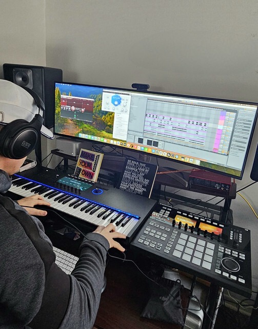 Music producer using a MIDI keyboard and software on dual monitors in a home studio, focused on creating audio tracks.