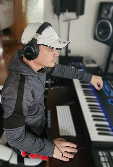 A musician wearing headphones and a hat focuses on a keyboard within a home studio setting, highlighting the creative process of music production.
